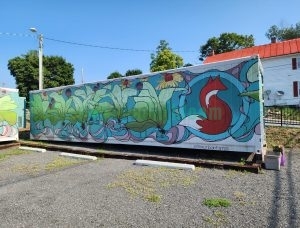 Blue shipping container farm side panel featuring painted red fox surrounded by green plants, butterflies, and pink water streams with #foxurbanfarms hashtag